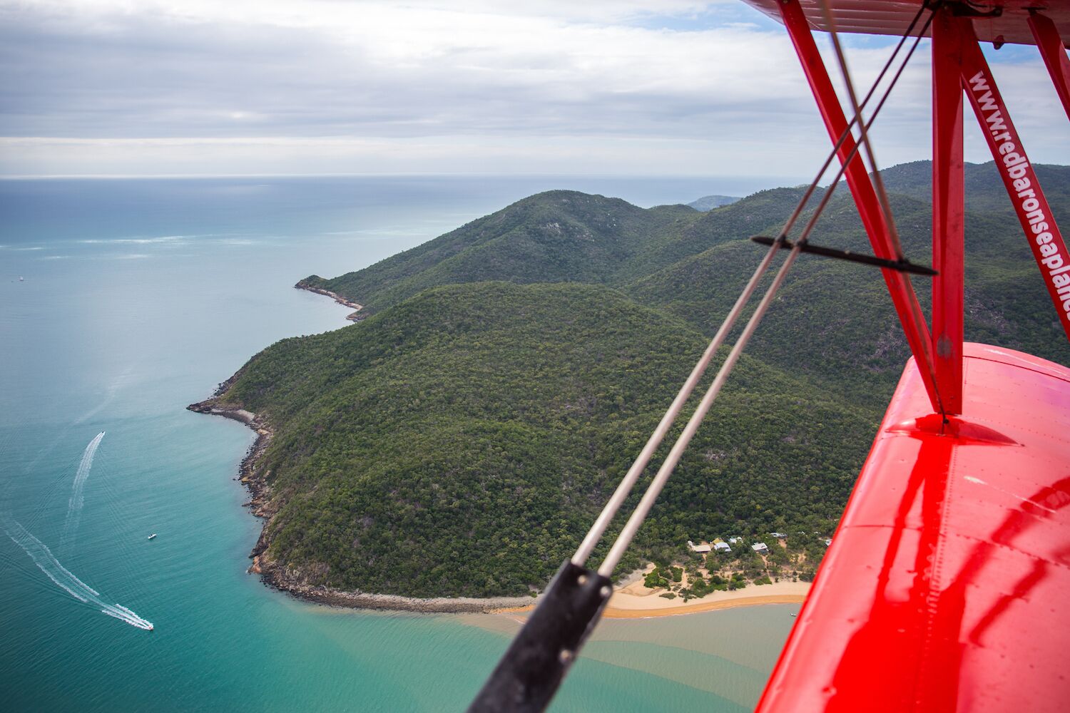 An aerial view of a coastal landscape with a beach, forested hills, and a clear blue sea.