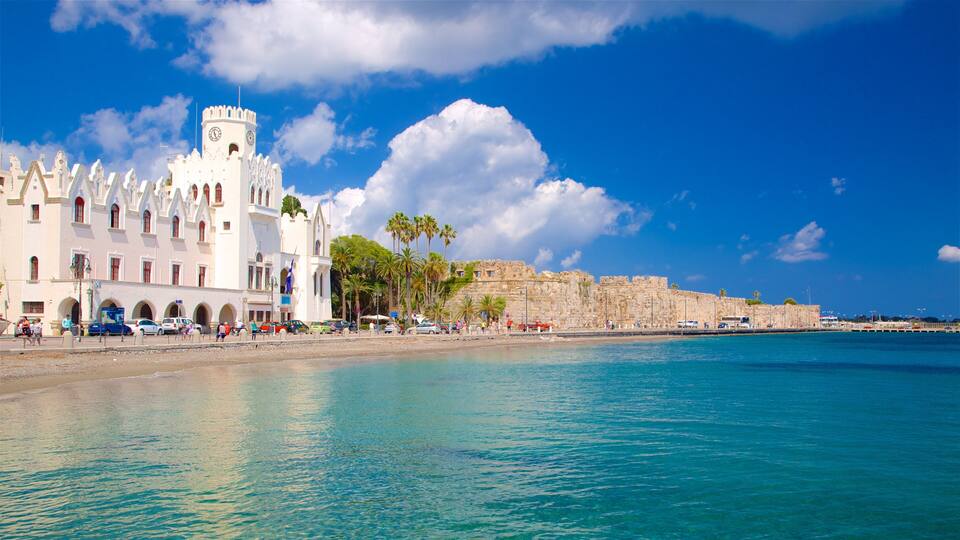 A coastal town with a prominent white building with a tower, palm trees, and a historic stone wall by the sea.