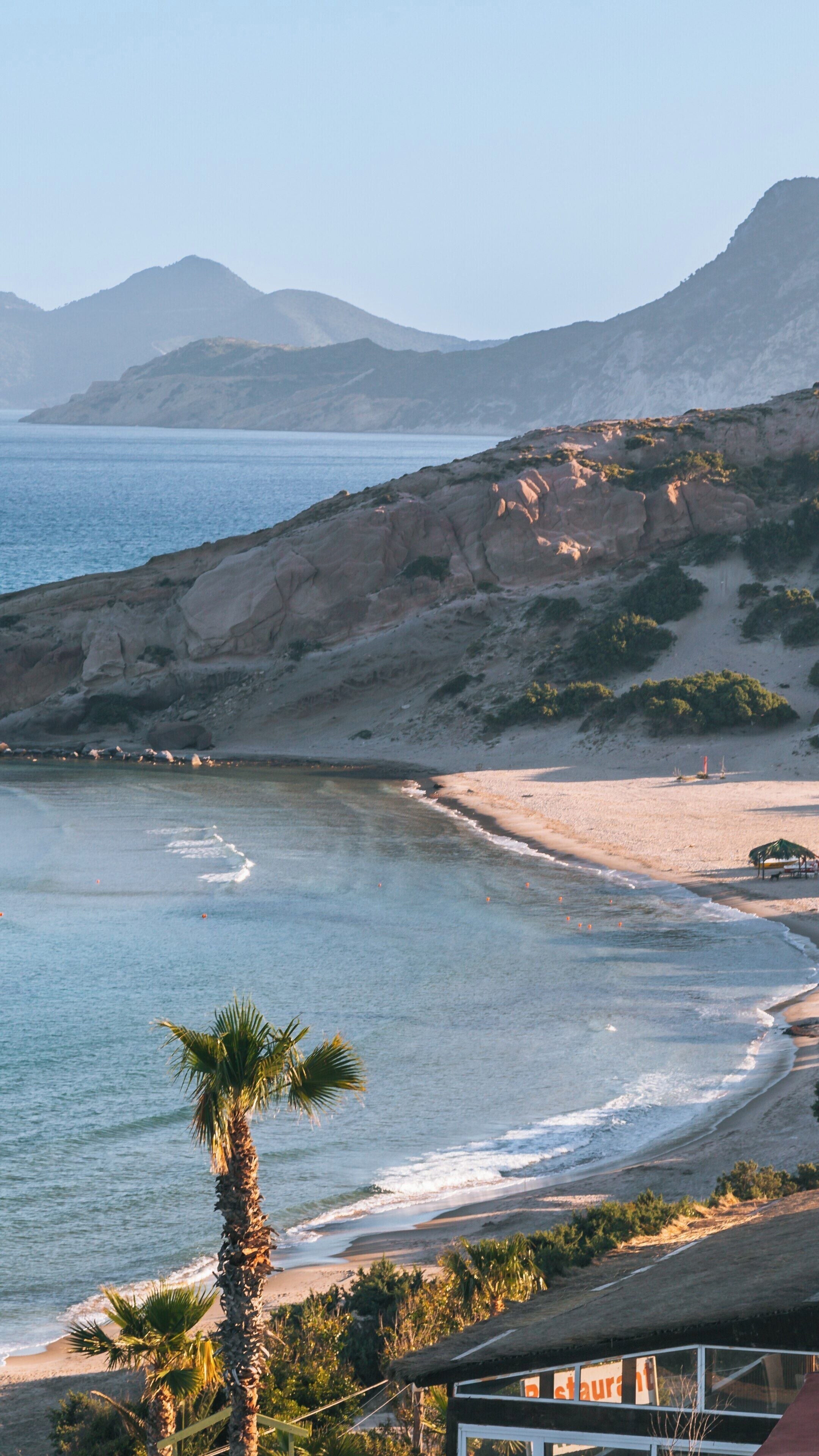 Stunning view of Paradise Beach in Kos, South Aegean, Greece, featuring crystal clear waters, gentle waves, and majestic mountains in the background during sunset