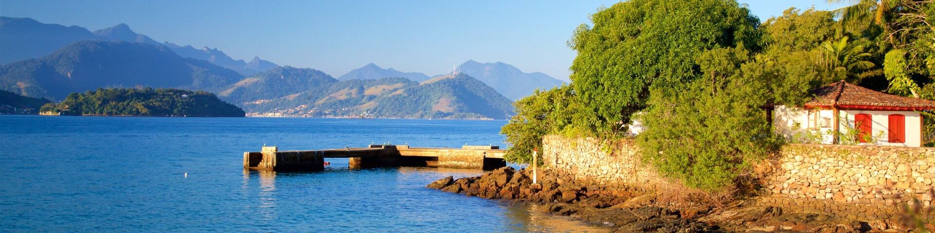 A coastal scene with a stone bridge over calm blue waters, a white building with red doors, and lush greenery.