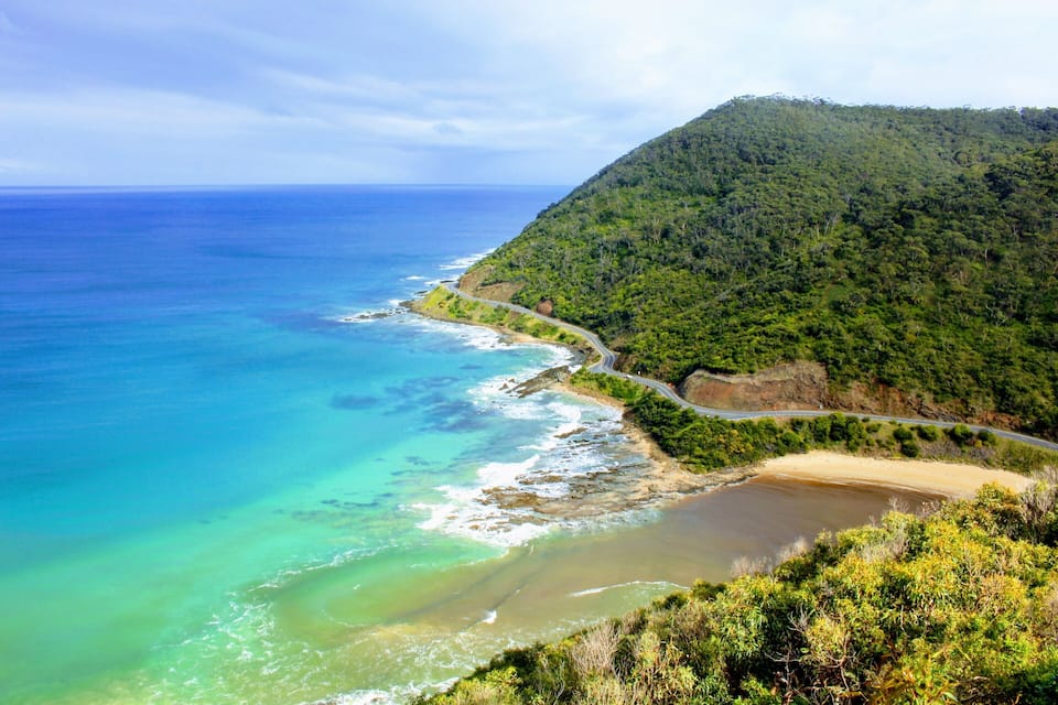 Un paisaje costero con un camino serpenteante a lo largo del acantilado, una playa y una colina boscosa.