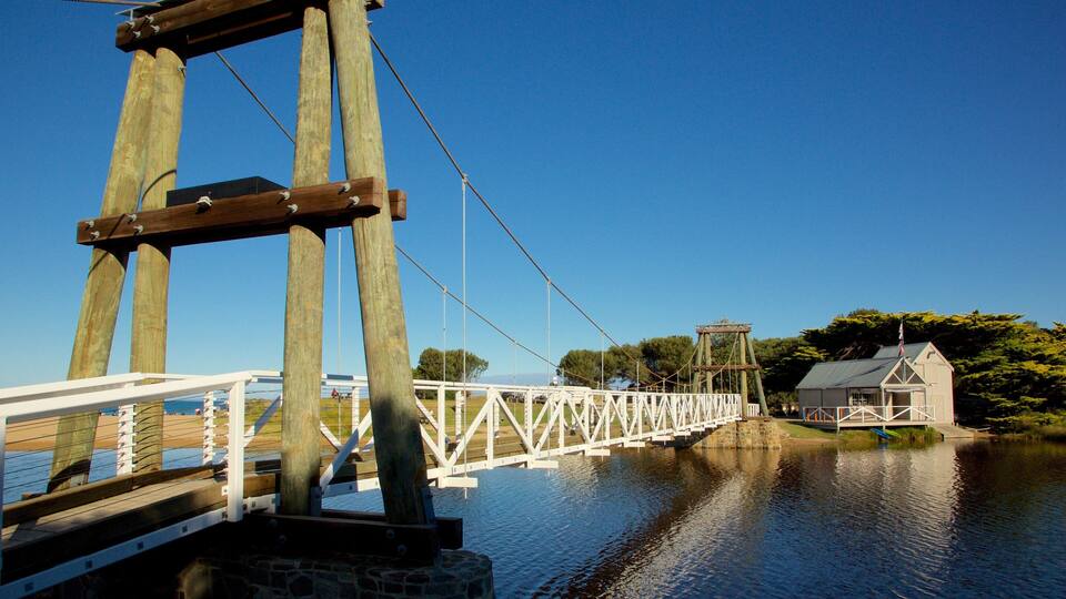 A wooden suspension bridge over a calm river, with a small building on the right bank.