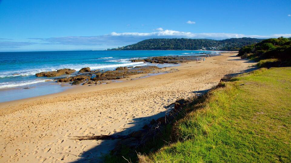 A sandy beach with rocky outcrops, a grassy area, and a distant hill with buildings.