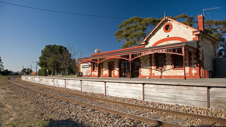 A historic train station with a distinctive red and white facade, surrounded by gravel and railway tracks.