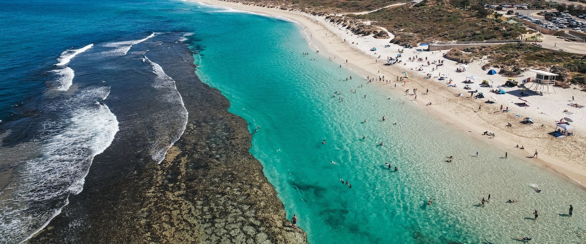 A coastal beach with clear turquoise waters, a sandy shore, and a few buildings in the background.