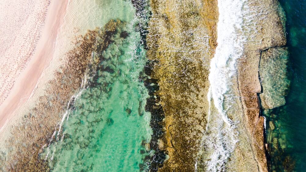 An aerial view of a beach with clear turquoise waters, a sandy shore, and rocky outcrops.