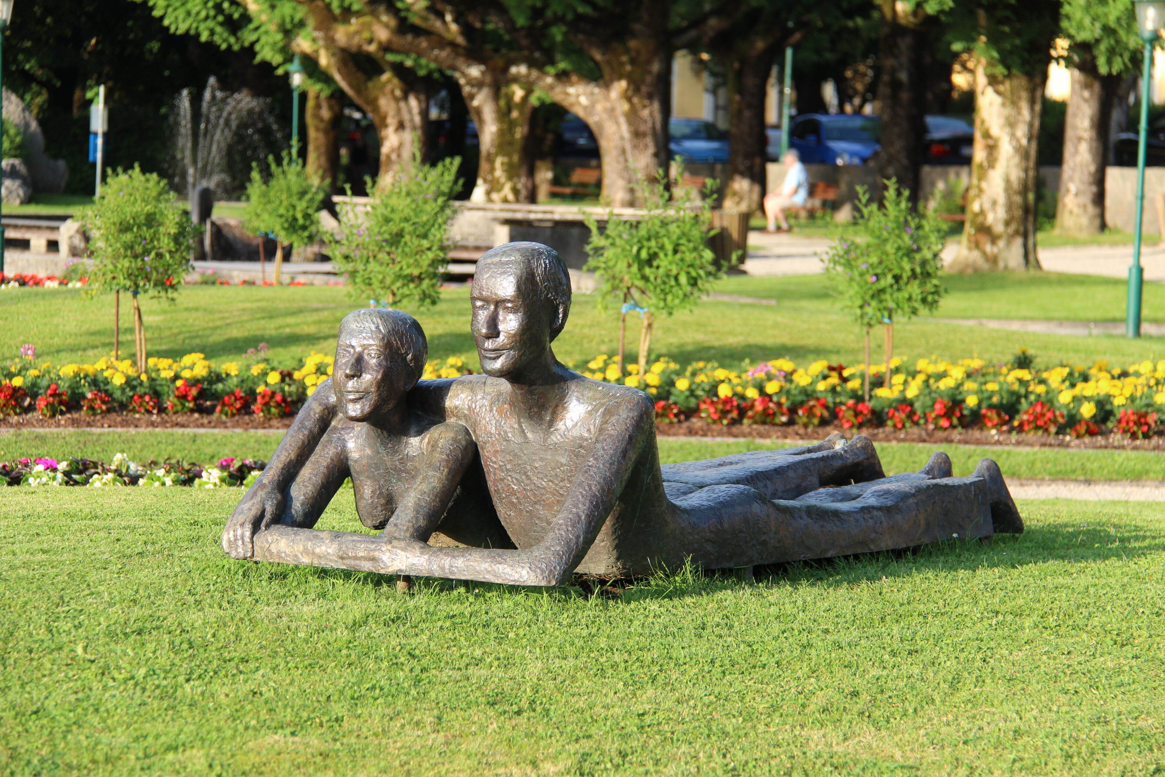 Escultura de duas figuras reclinadas numa área gramada, com canteiros de flores e árvores ao fundo.