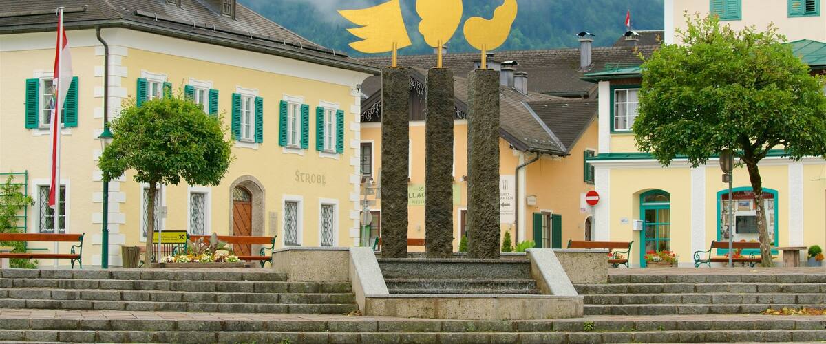 A town square with a fountain, benches, and buildings with green shutters.