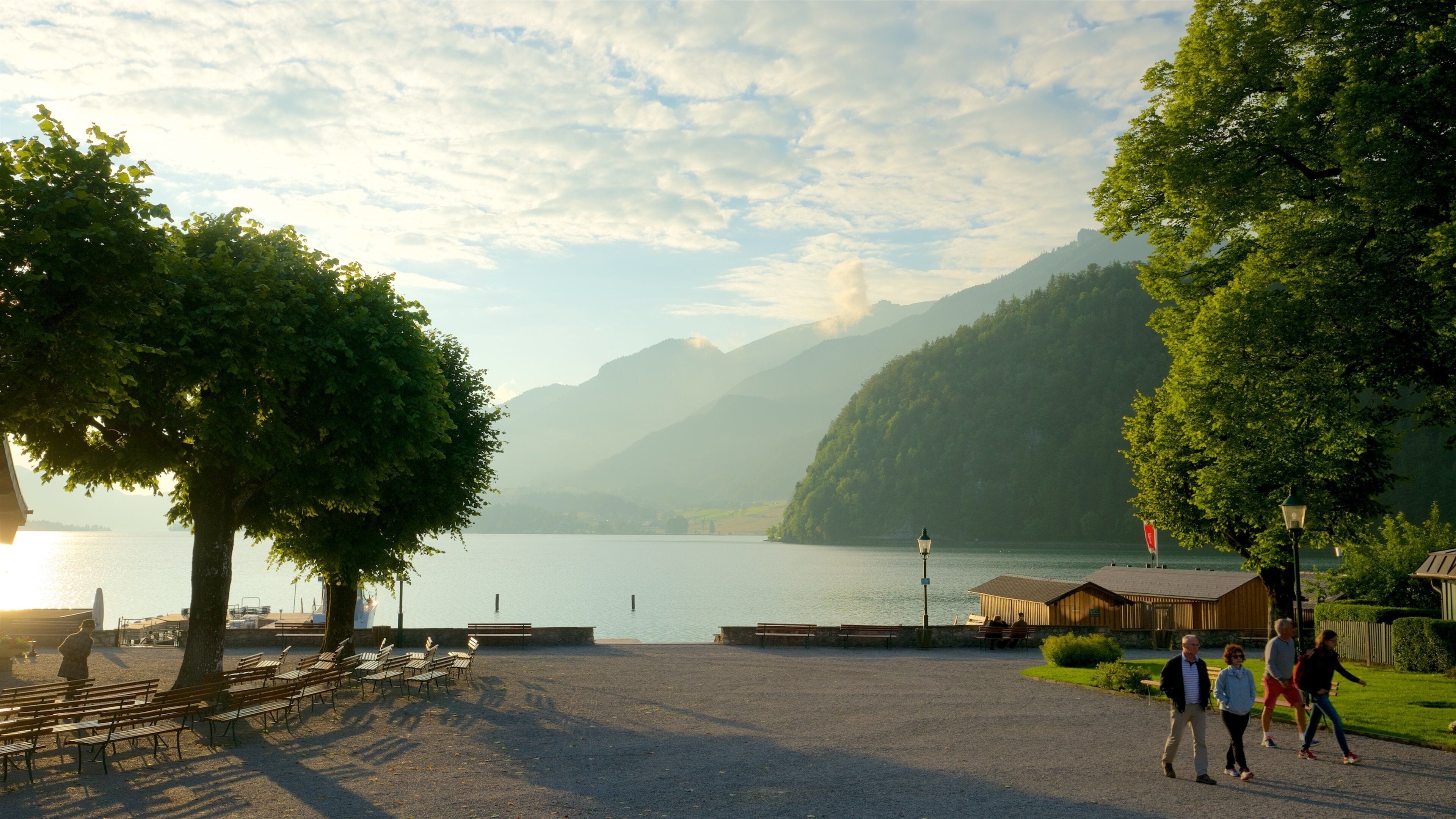 A lakeside park with benches, a wooden building, and mountains in the background.