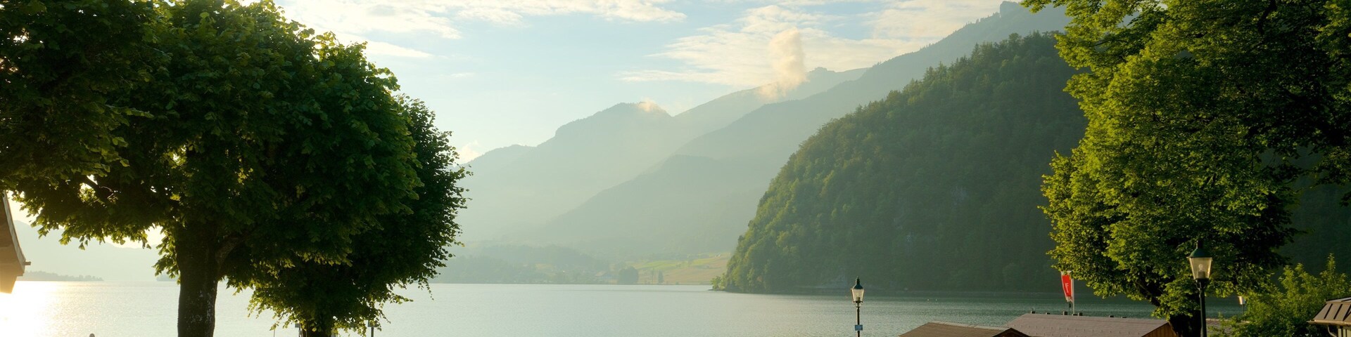 A lakeside park with benches, a wooden building, and mountains in the background.