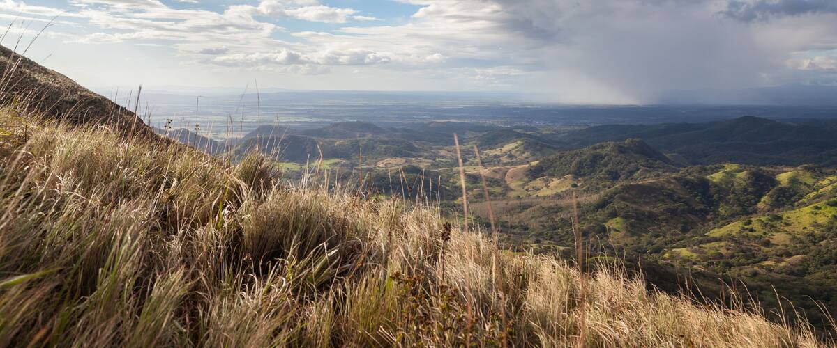 A grassy hillside with a panoramic view of rolling hills and distant cityscape under a partly cloudy sky.