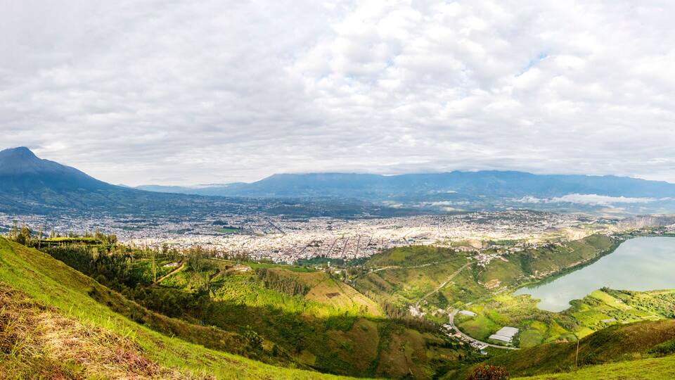 A panoramic view of a city with a large lake, surrounded by green hills and mountains.