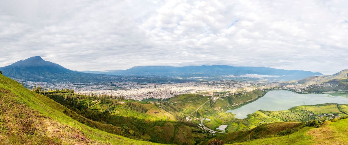 A panoramic view of a city with a large lake, surrounded by green hills and mountains.
