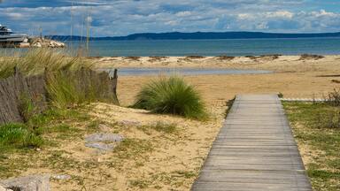 Une promenade en bois mène à une plage de sable fin, bordée par une mer calme et dominée par des montagnes lointaines.