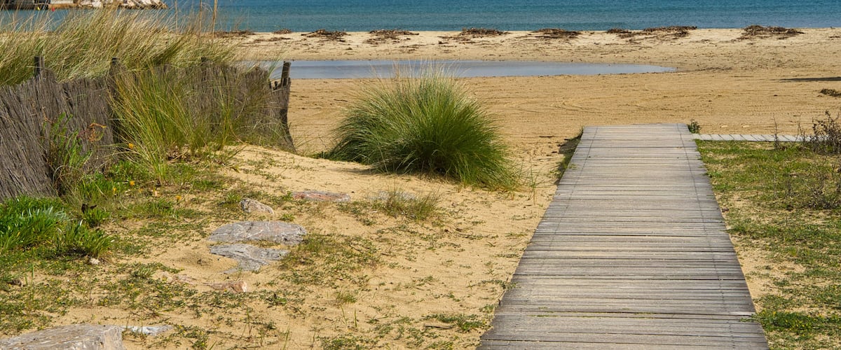 A wooden boardwalk leading to a sandy beach with a calm sea and distant mountains.