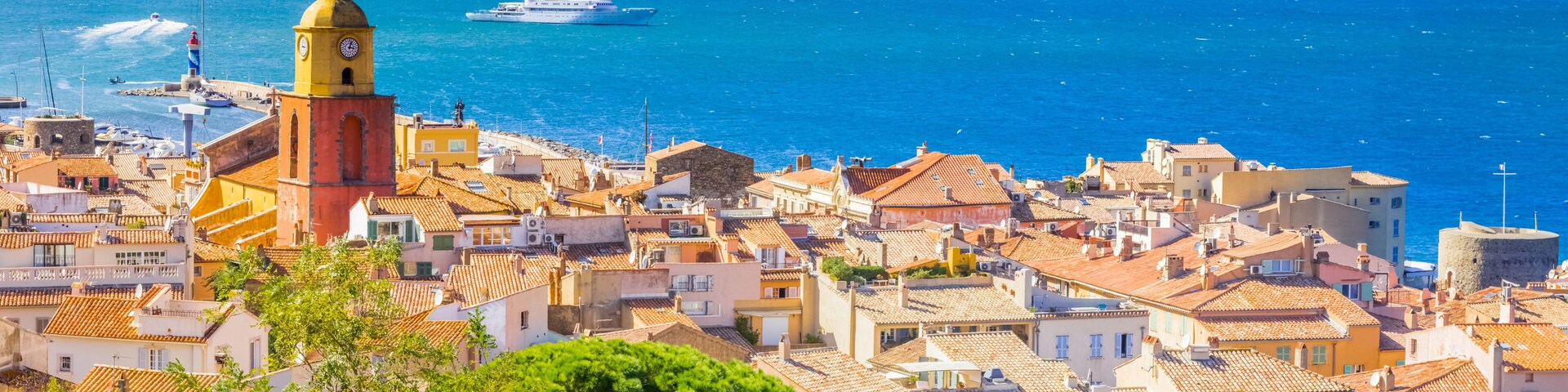 A coastal town with terracotta-roofed buildings, a lighthouse, and a cruise ship in the background.
