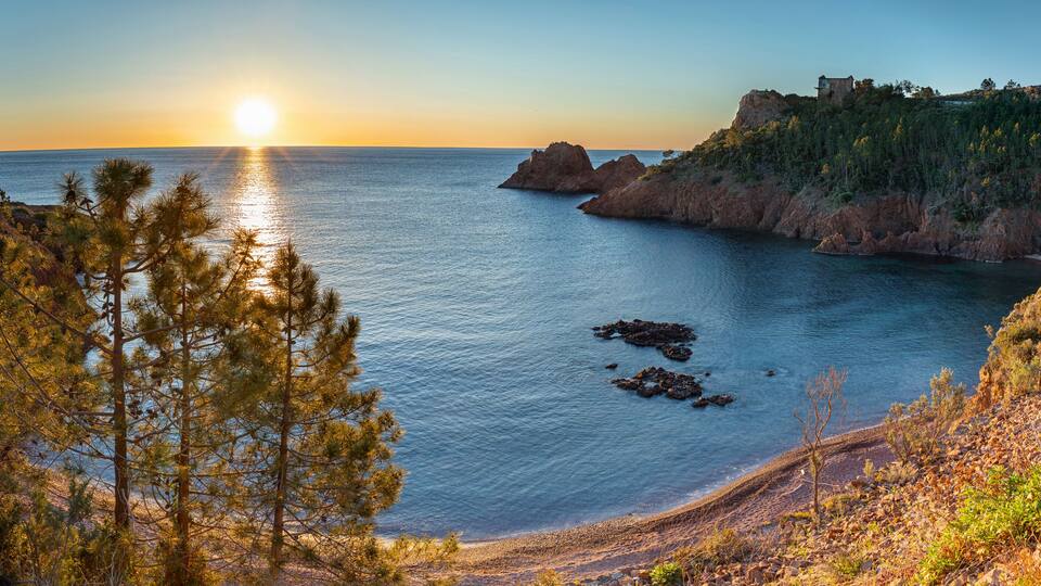 A coastal landscape with a sunset, rocky cliffs, and a small building on a hill.