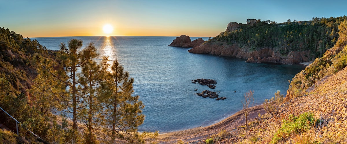 A coastal landscape with a sunset, rocky cliffs, and a small building on a hill.