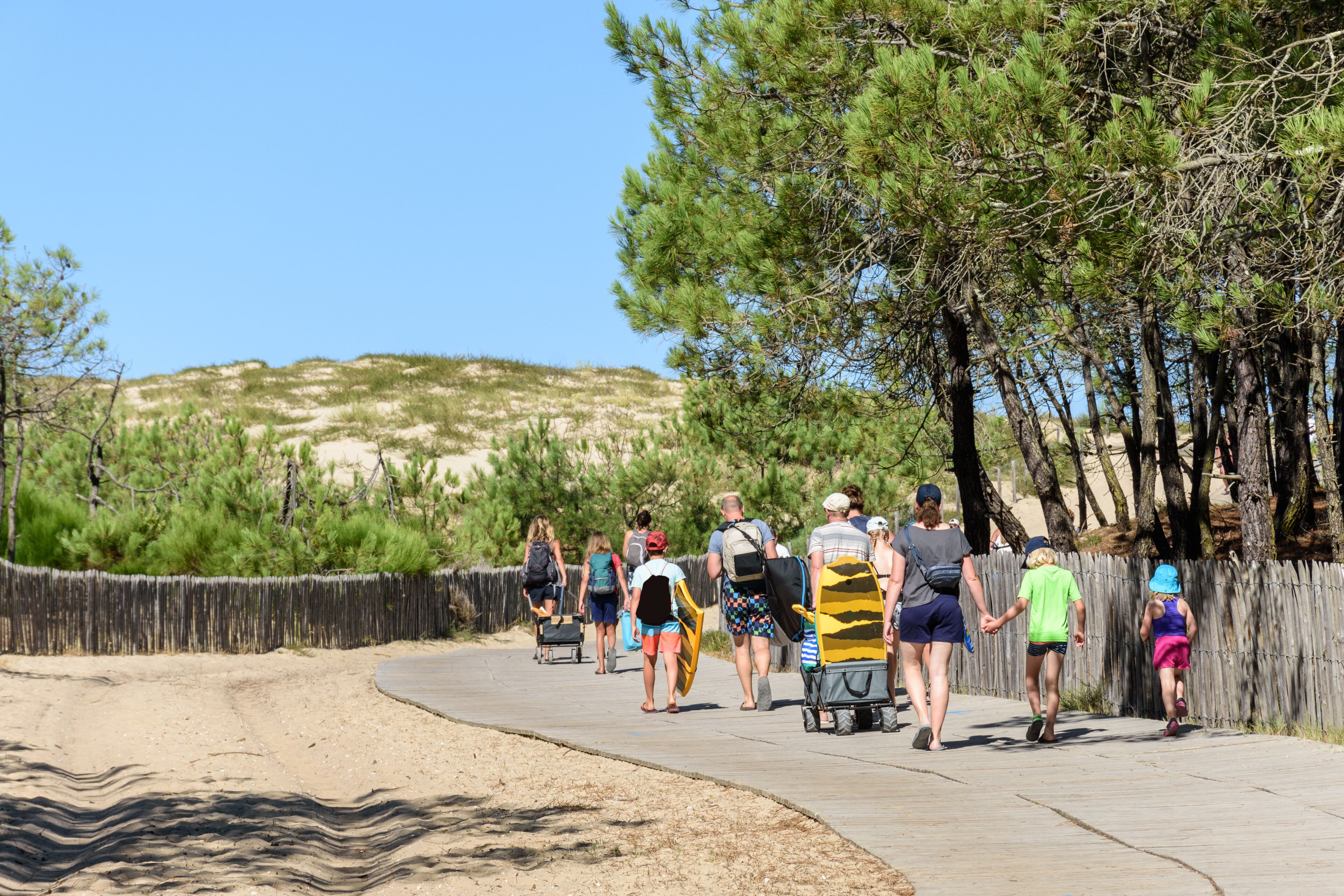 A sandy beach with a wooden fence, a clear blue sky, and a group of people walking along a boardwalk.