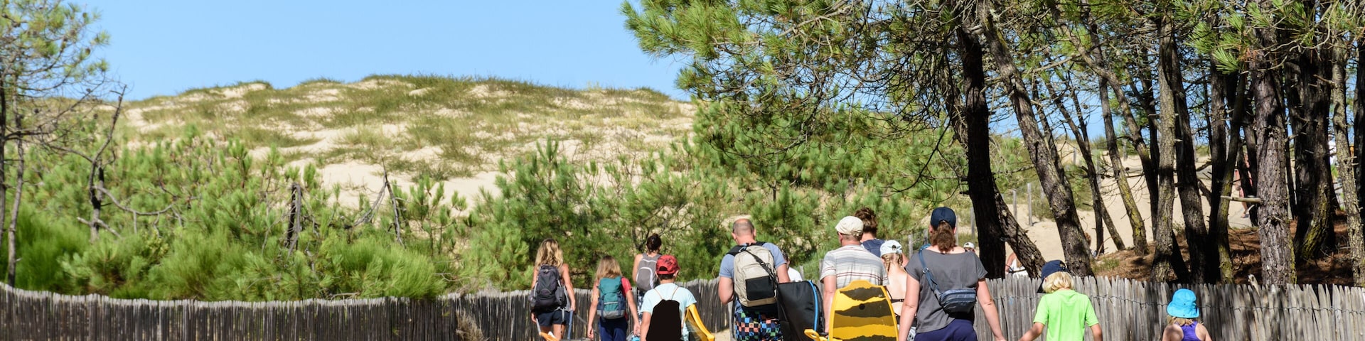 A sandy beach with a wooden fence, a clear blue sky, and a group of people walking along a boardwalk.