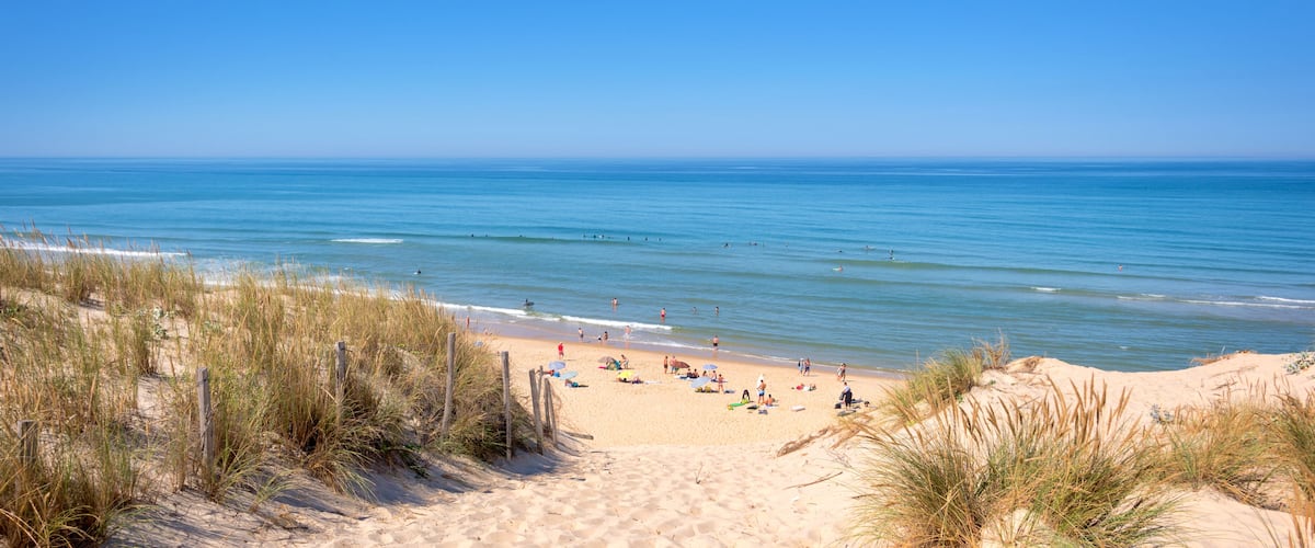 A sandy beach with people sunbathing and swimming, surrounded by dunes and a clear blue ocean.