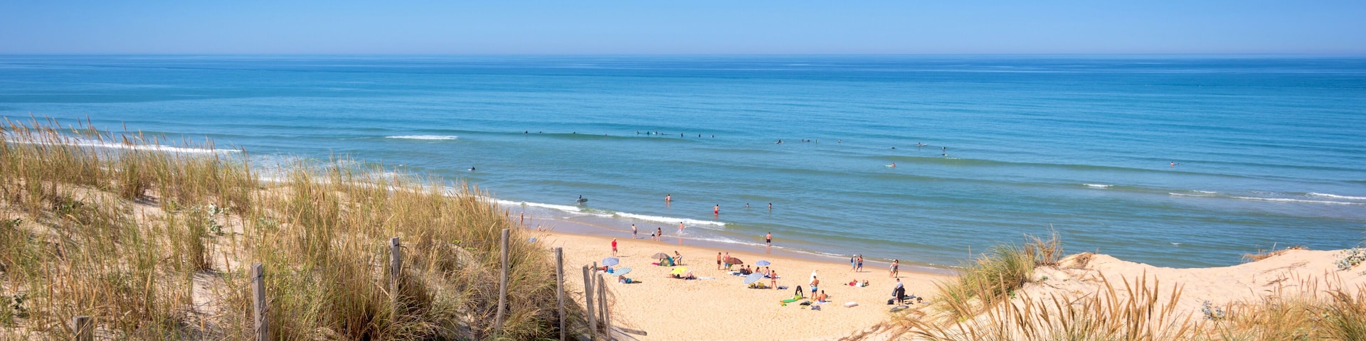 A sandy beach with people sunbathing and swimming, surrounded by dunes and a clear blue ocean.