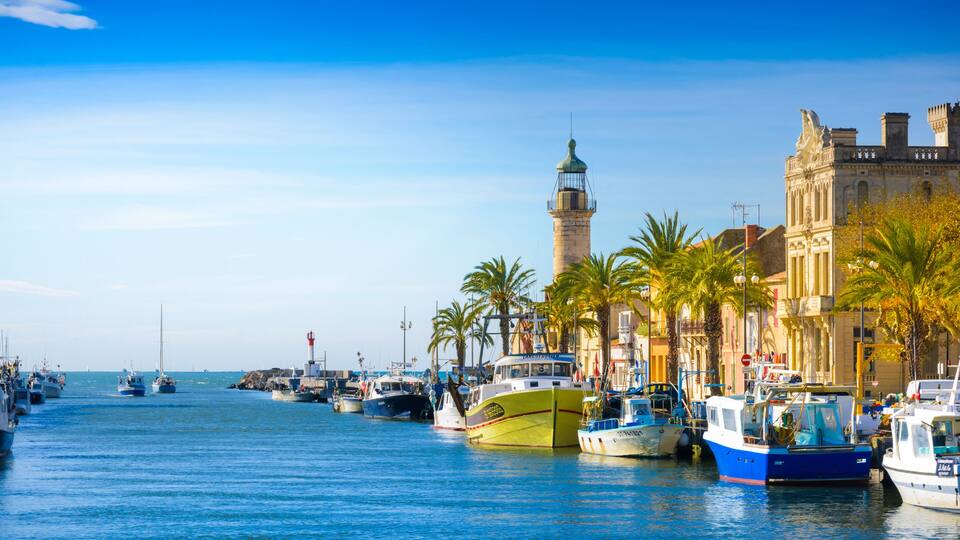 A harbor with boats, a lighthouse, and a historic building with palm trees.