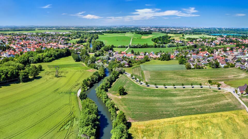 An aerial view of a town with a river, green fields, and red-roofed houses.