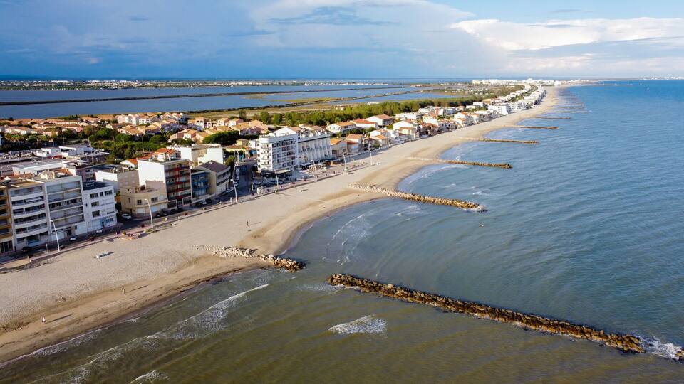 A coastal cityscape with a long sandy beach, a rocky breakwater, and residential buildings along the shore.