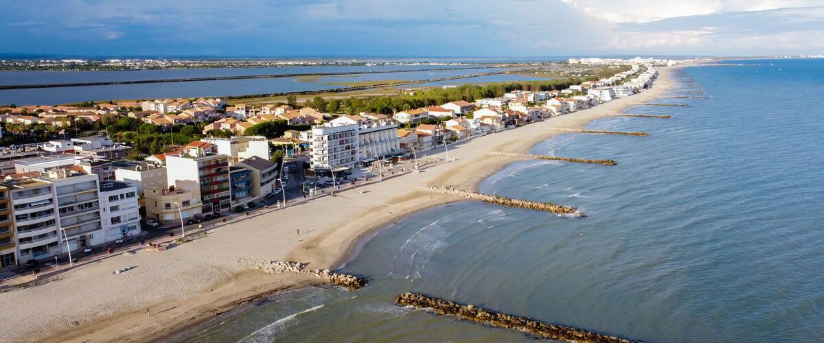 A coastal cityscape with a long sandy beach, a rocky breakwater, and residential buildings along the shore.