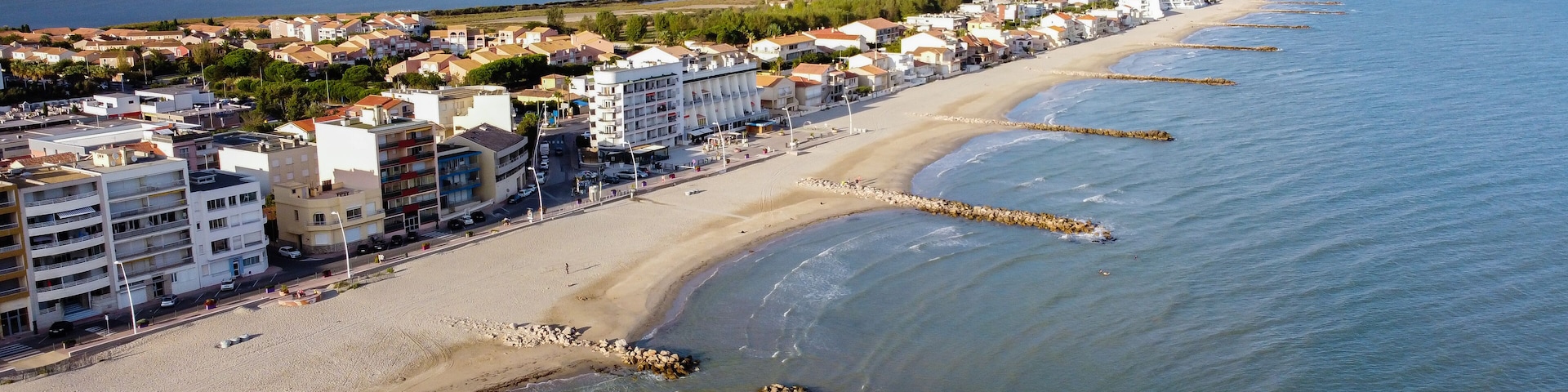 A coastal cityscape with a long sandy beach, a rocky breakwater, and residential buildings along the shore.