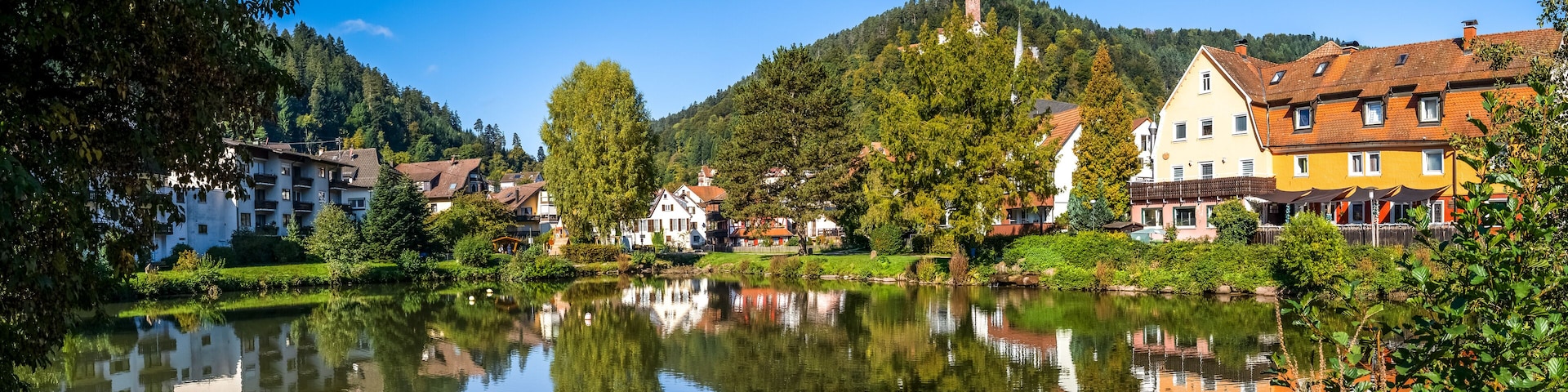 Um lago calmo com reflexos de edifícios e árvores, rodeado por vegetação exuberante e uma colina com uma torre.