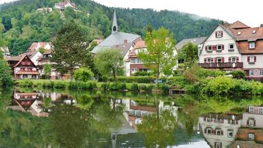 A calm river reflecting buildings and a church steeple on a hillside.