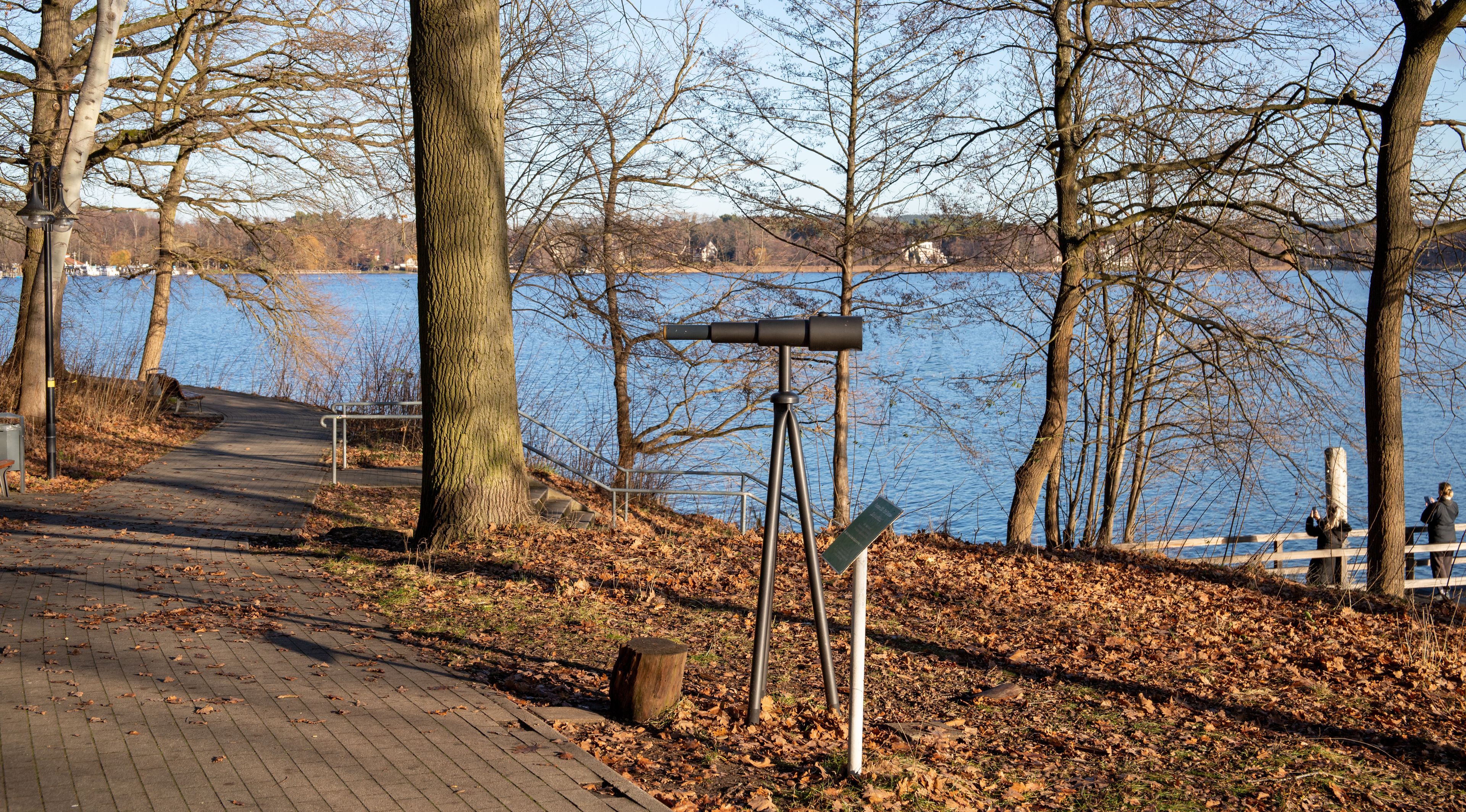 A lakeside park with a walking path, leaf-covered ground, and a telescope on a tripod.