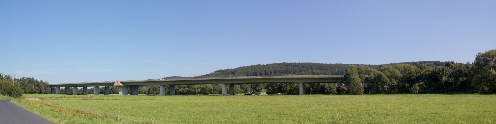 A highway overpass with a red triangular warning sign, surrounded by green fields and a forested hill.