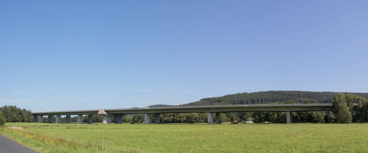 A highway overpass with a red triangular warning sign, surrounded by green fields and a forested hill.