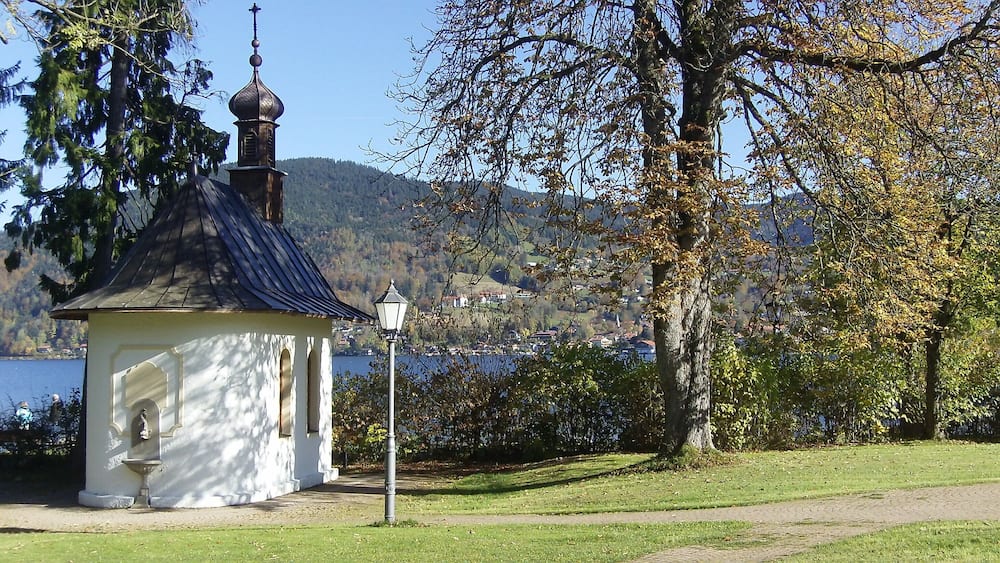 A small white chapel with a dark roof and a bell tower, surrounded by trees and a lake.