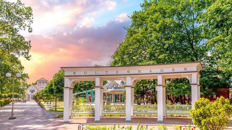 A white pavilion with 'BOLTENHAGEN' inscribed, surrounded by a garden with various flowers and trees.