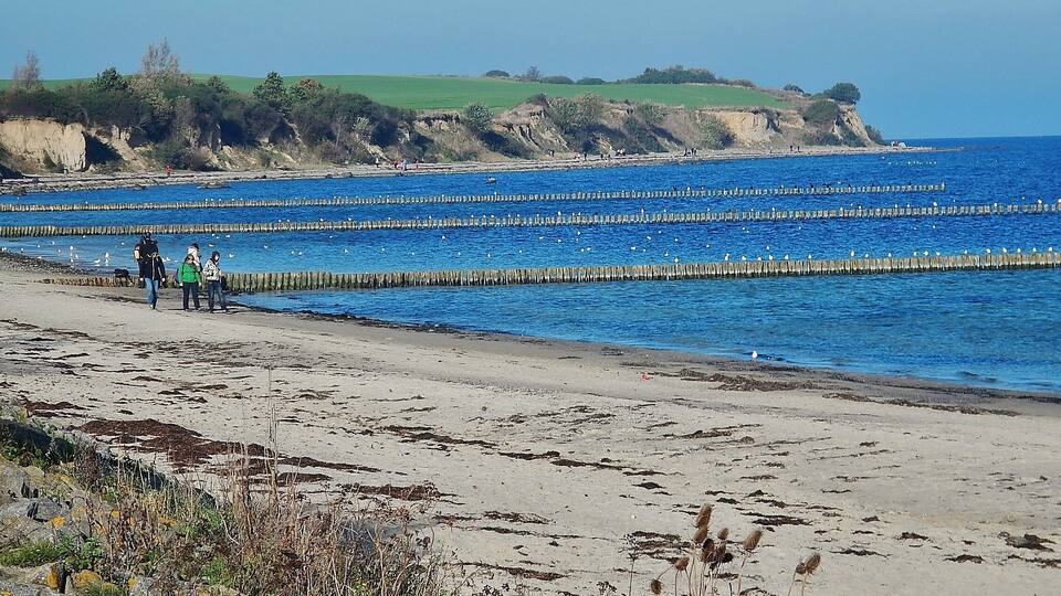 A beach with a clear blue sea, a rocky cliff, and a grassy hill in the background.