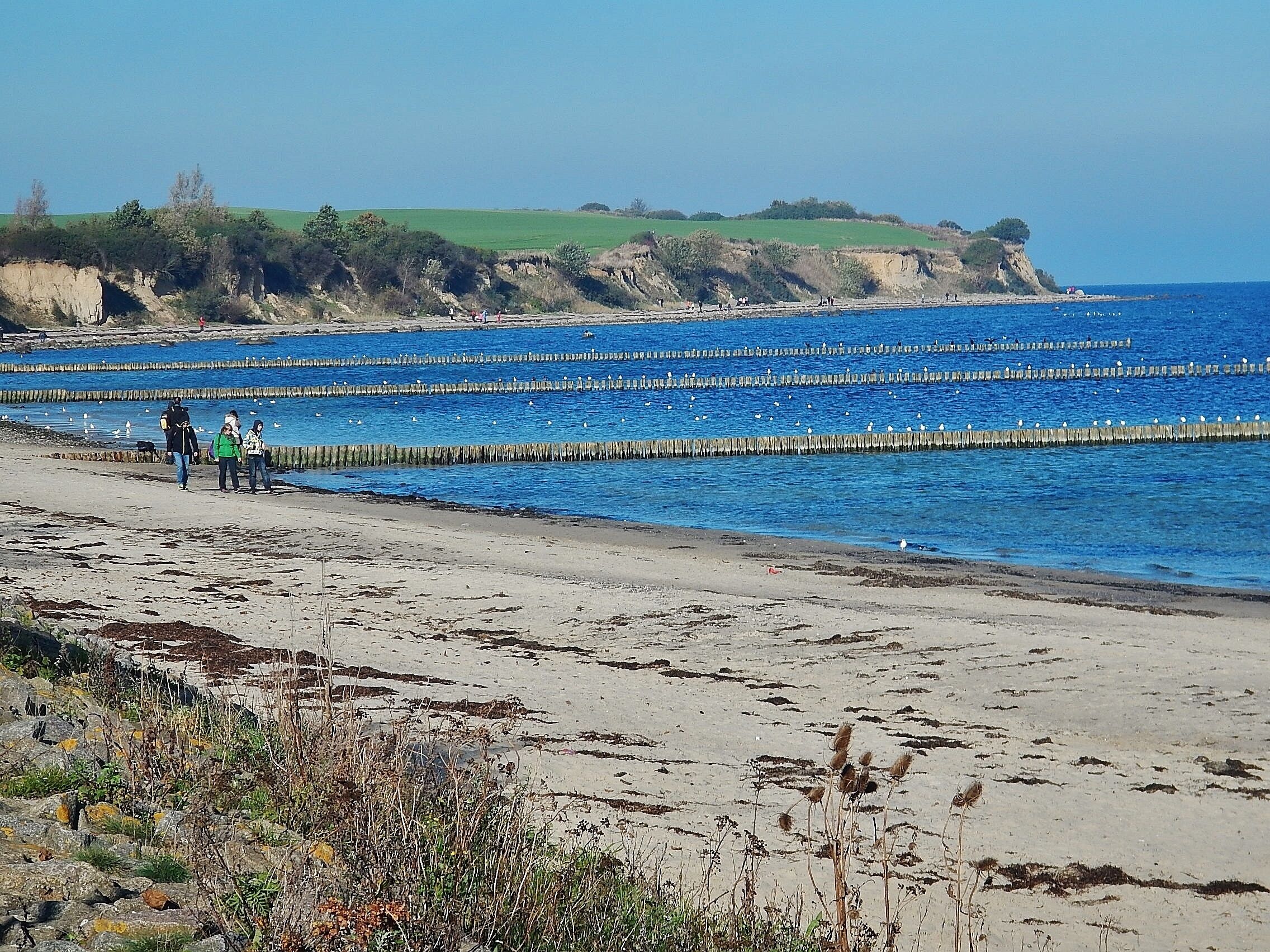 A beach with a clear blue sea, a rocky cliff, and a grassy hill in the background.