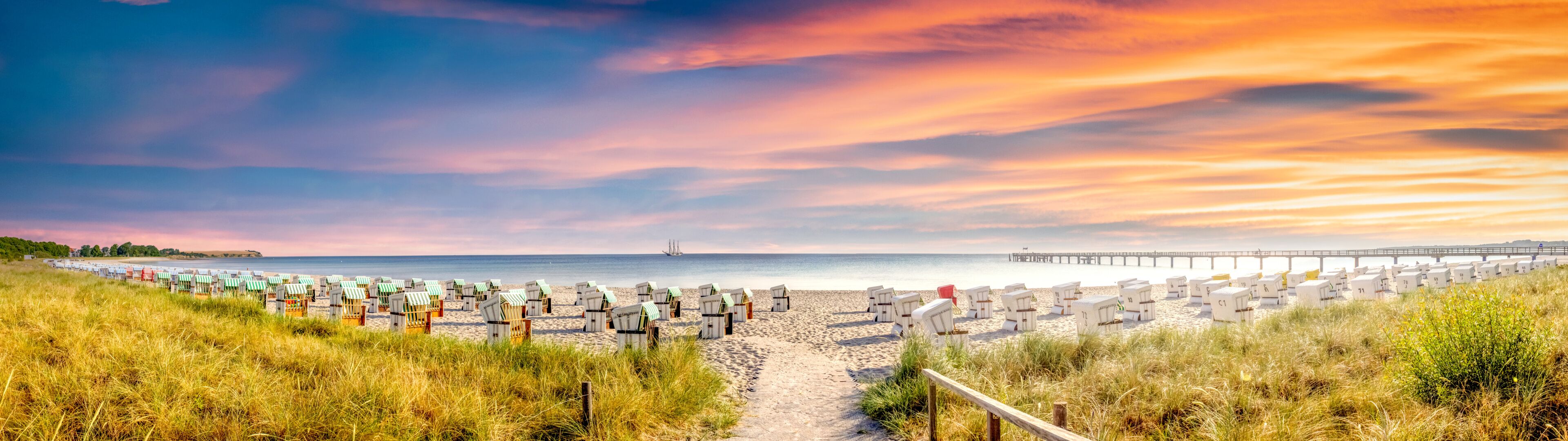 A beach with rows of beach huts, a pier extending into the water, and a clear sky with clouds.