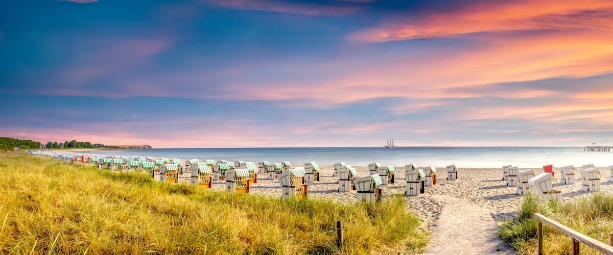 A beach with rows of beach huts, a pier extending into the water, and a clear sky with clouds.
