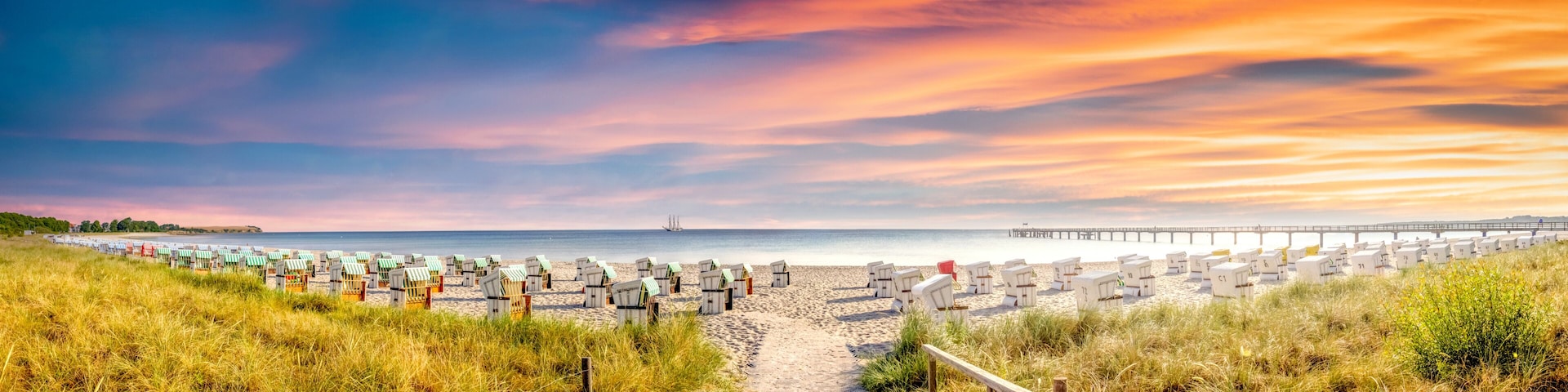 A beach with rows of beach huts, a pier extending into the water, and a clear sky with clouds.
