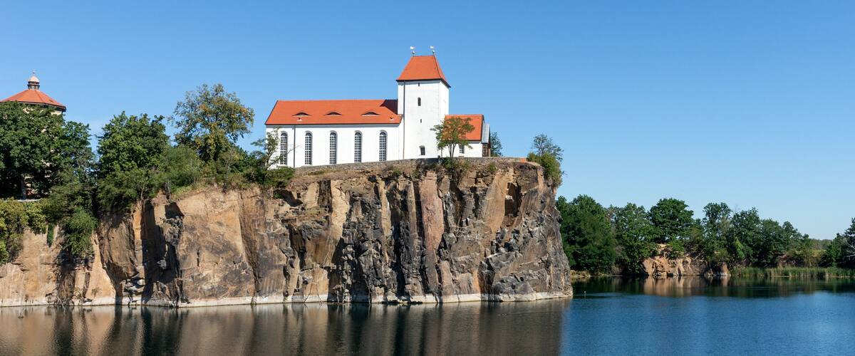 A castle on a cliff overlooking a calm lake, surrounded by trees and rocky cliffs.