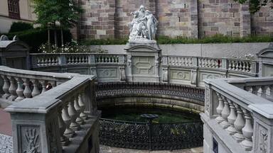 Une fontaine classique ornée d’une statue, entourée de balustrades en pierre travaillées et d’un mur de pierre.