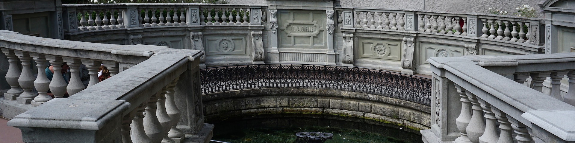 A classical fountain with a statue, surrounded by ornate stone balustrades and a stone wall.