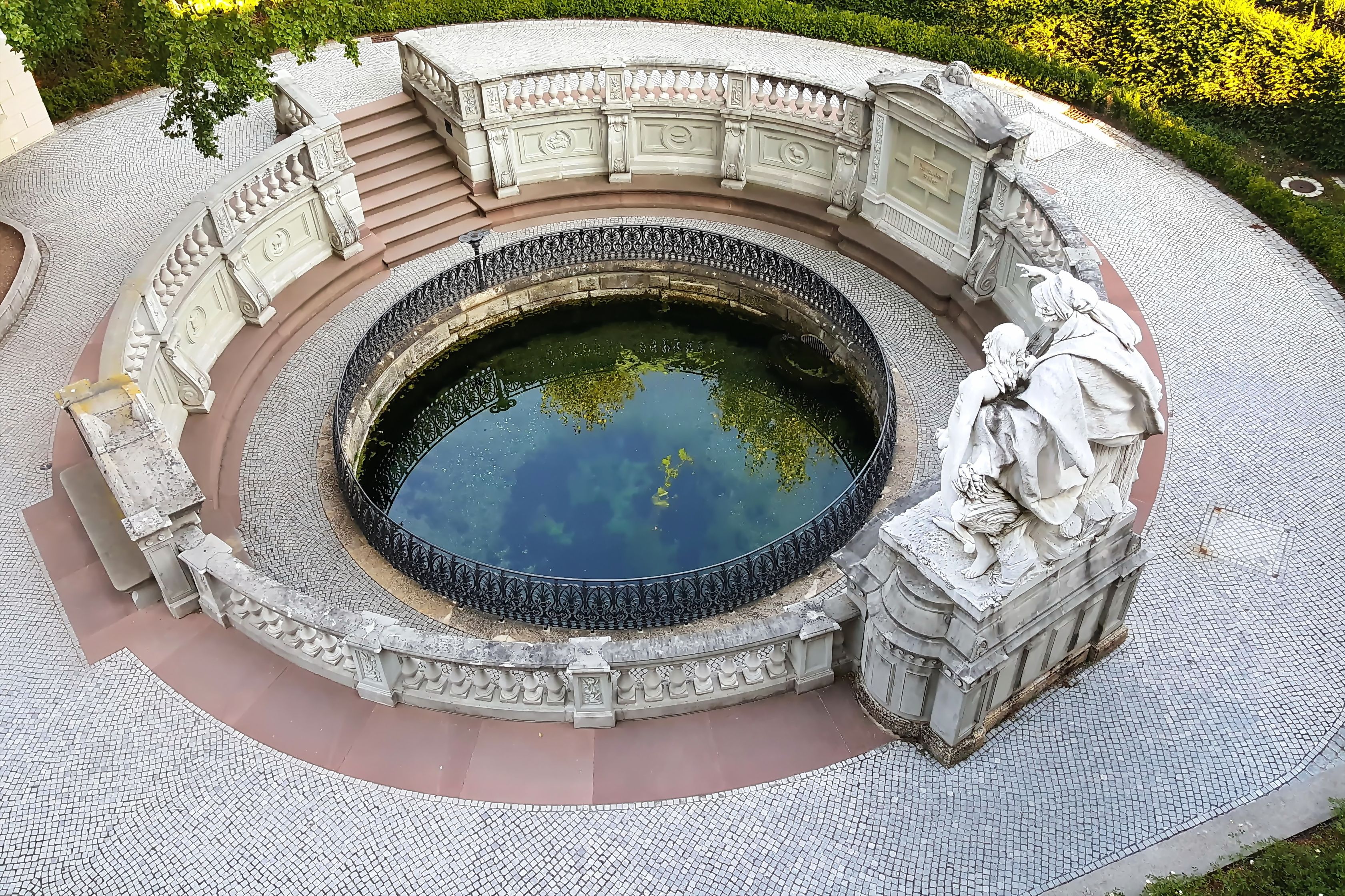 Een ronde fontein met een standbeeld, omgeven door versierde stenen balustrades en een betegeld voetpad.