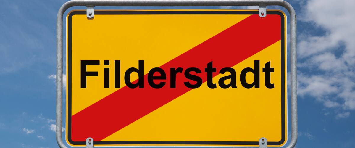 A yellow sign with 'Filderstadt' written on it, mounted on a metal pole against a blue sky with clouds.