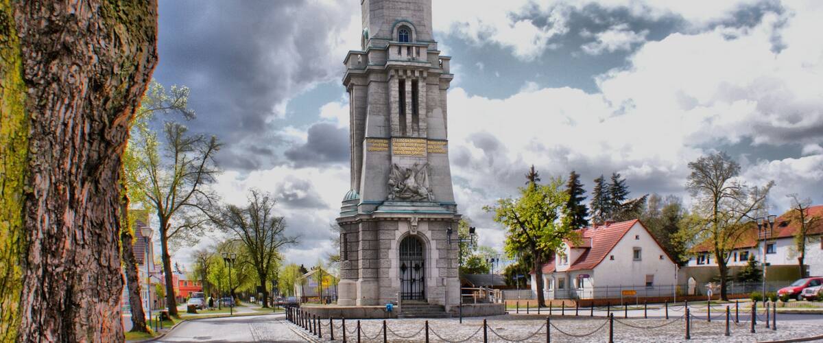 A tall stone monument with inscriptions and a clock, surrounded by trees and buildings.