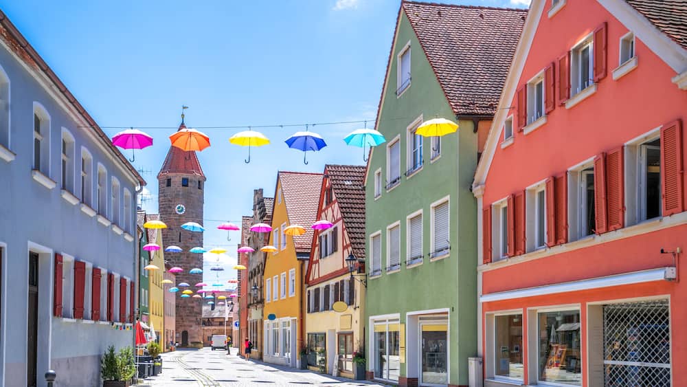 A cobblestone street lined with colorful buildings, a tower, and string lights with umbrellas.